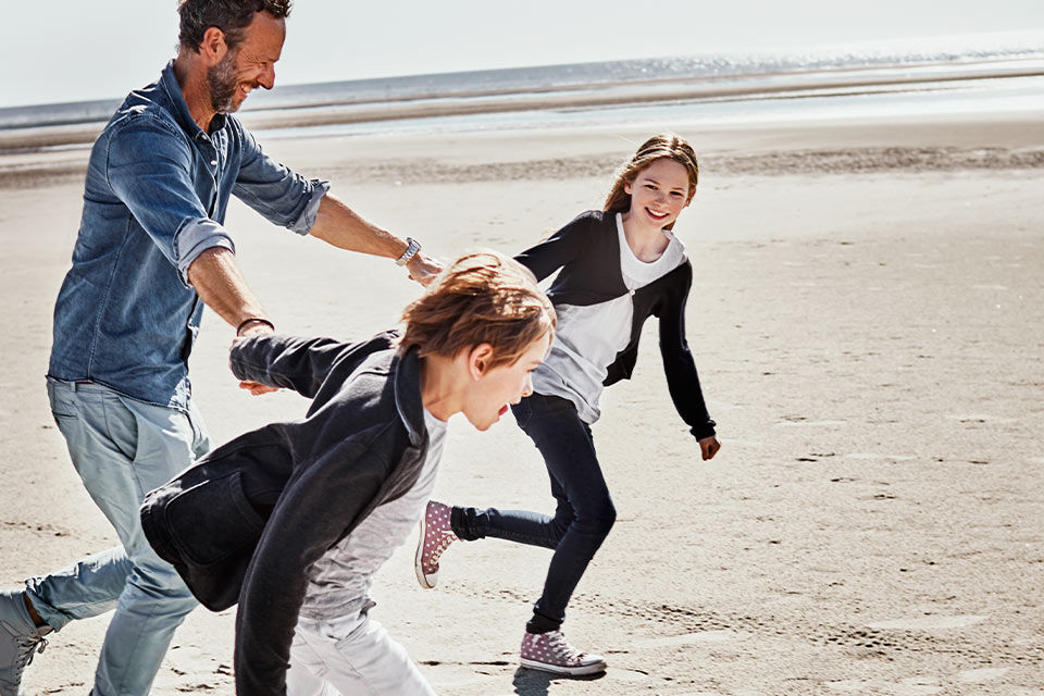 Ein Mann spielt mit zwei Kindern am Strand; alle laufen lachend über den Sand.