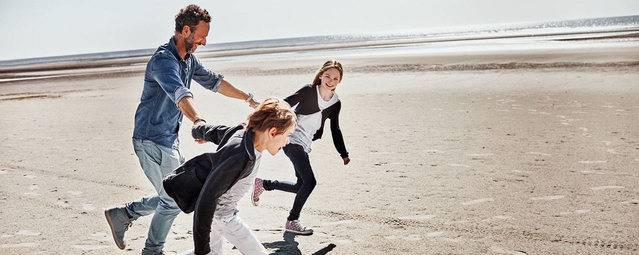 Ein Mann spielt mit zwei Kindern am Strand; alle laufen lachend über den Sand.