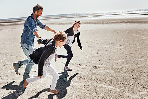 Zwei Kinder ziehen ihren Vater an den Händen an einem Strand.