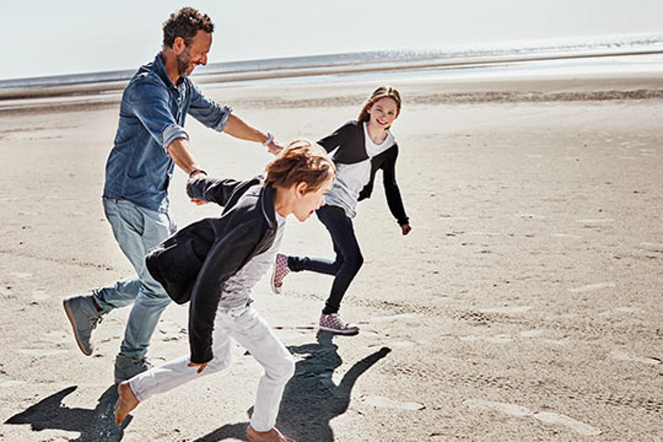 Ein Vater läuft mit seinen Kindern an der Hand lächelnd an einem Strand entlang.