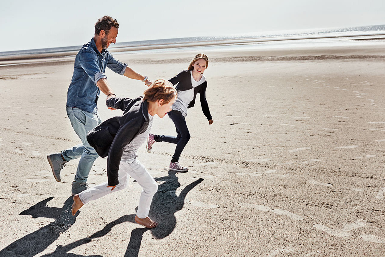 Ein Mann spielt mit zwei Kindern am Strand; alle laufen lachend über den Sand.