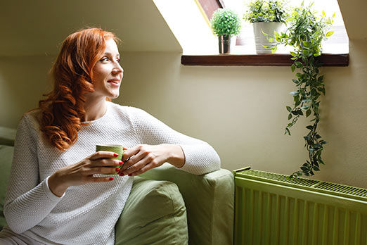 Frau mit Kaffeetasse in der Hand sitzt auf dem Sofa neben der Heizung und schaut aus dem Fenster