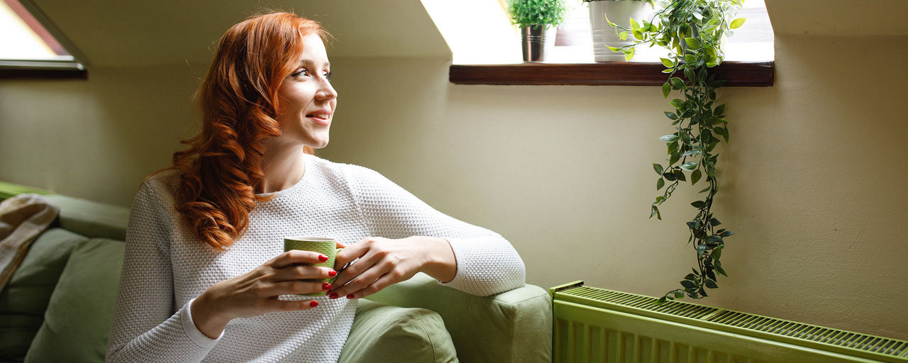 Frau mit Kaffeetasse in der Hand sitzt auf dem Sofa neben der Heizung und schaut aus dem Fenster