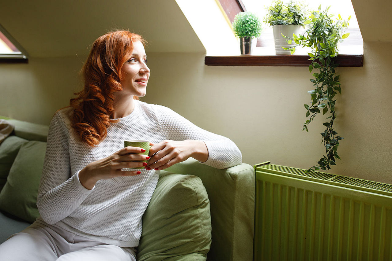 Eine Frau sitzt auf einem grünen Sofa in einer Dachwohnung und sieht zum Dachfenster hinaus.