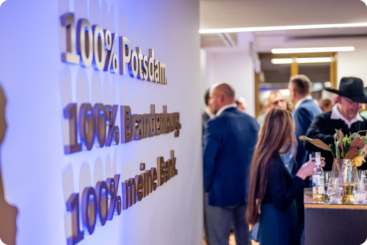 Im Vordergrund steht ein Bild mit der Aufschrift 100% Potsdam, 100% Brandenburg und 100% meine Bank und im Hintergrund stehen Menschen