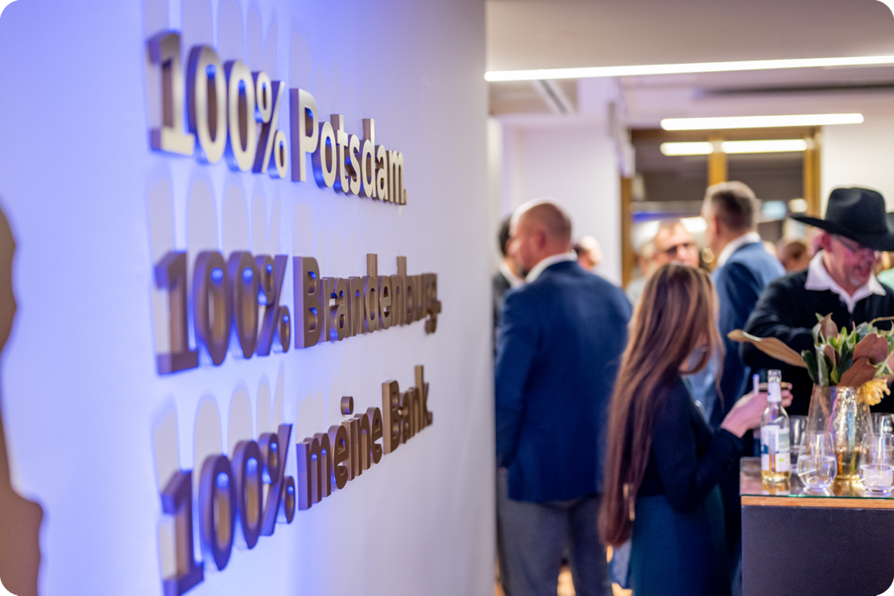 Im Vordergrund steht ein Bild mit der Aufschrift 100% Potsdam, 100% Brandenburg und 100% meine Bank und im Hintergrund stehen Menschen