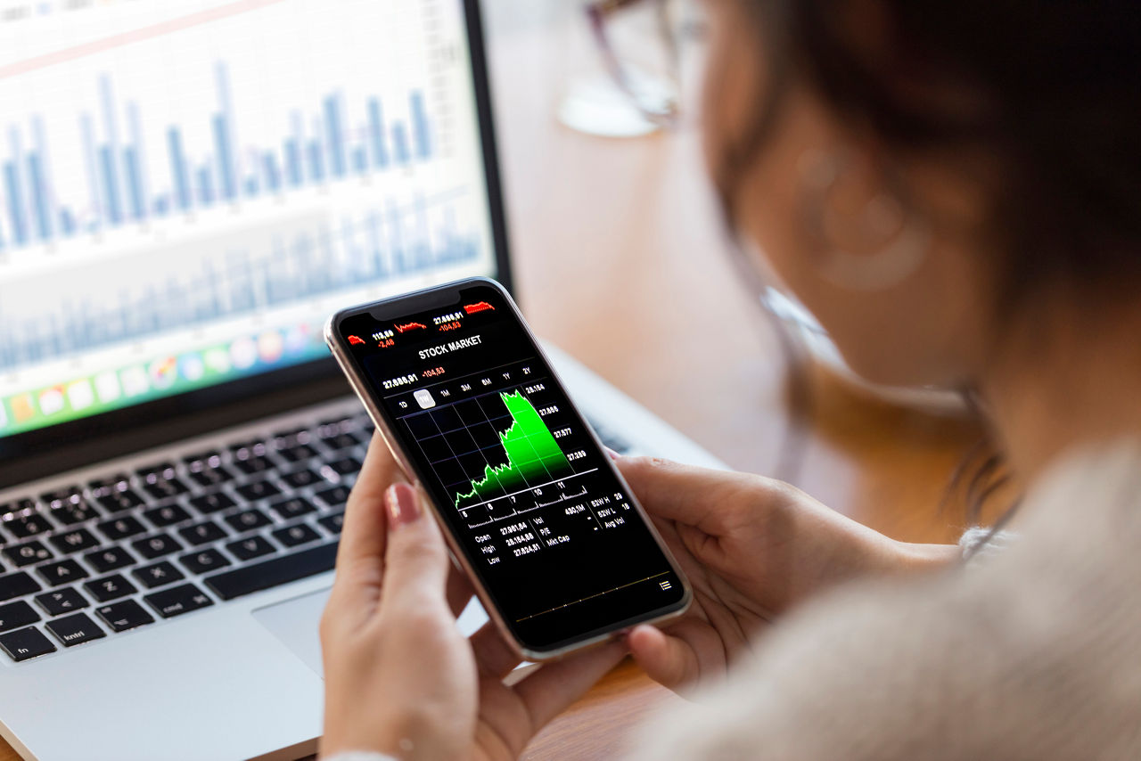 Stock Market wird auf Handy angezeigt, im Hintergrund steht ein Laptop