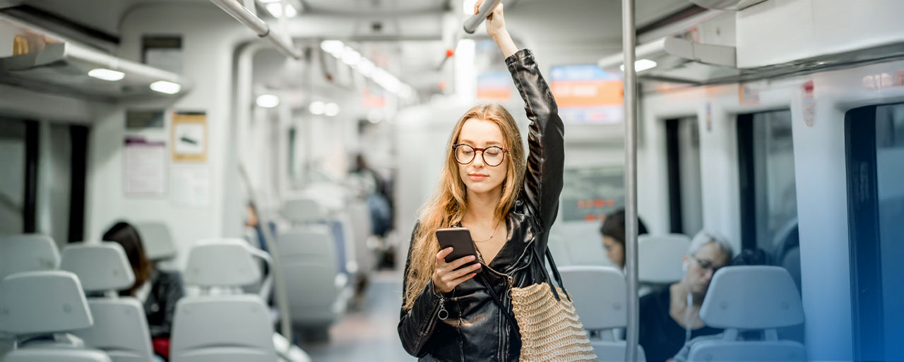 Junge Frau im Zug stehend, hält sich oben an der Stange fest und blickt auf Ihr Smartphone