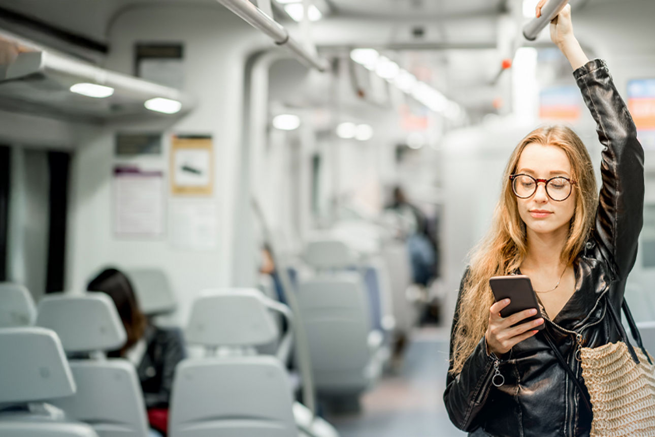 Junge Frau im Zug stehend, hält sich oben an der Stange fest und blickt auf Ihr Smartphone