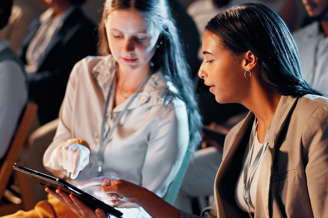 Zwei junge Frauen sitzen im Publikum nebeneinander und zeigen sowie schauen auf ein Tablet