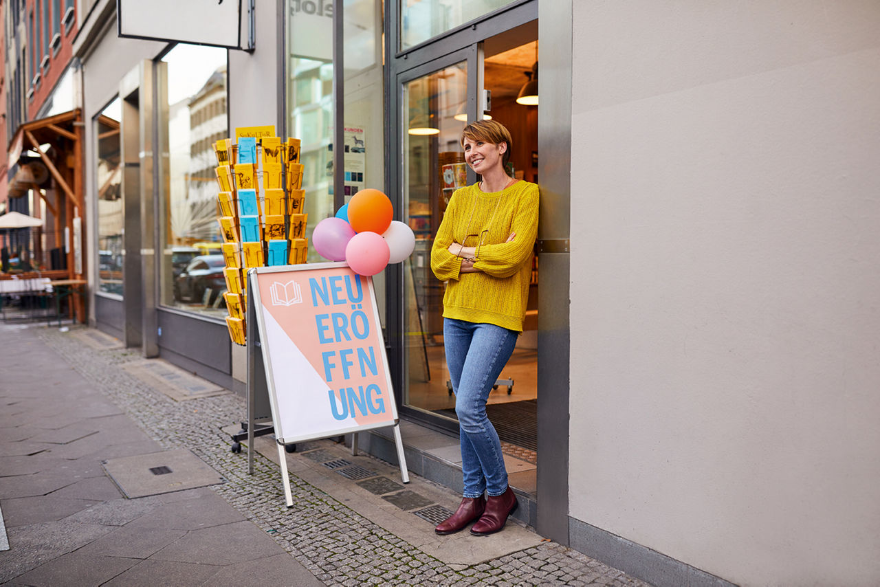 Junge Frau steht vor ihrem Geschäft, der Aufsteller zur Neueröffnung ist mit Luftballons geschmückt.
