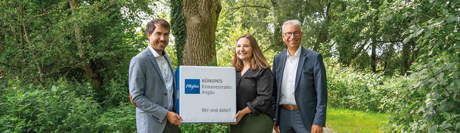 Ein Mann und eine Frau halten einen Würfel in die Höhe mit der Aufschrift "Bündnis klimaneutrales Allgäu. Wir sind dabei!". Daneben steht ein weiterer Mann. Sie stehen im Grünen.