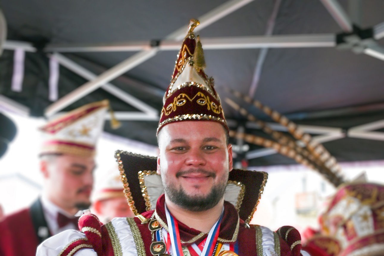 Prinz Hasi mit rot-weißem Gewand, Narrenkappe und vielen Karnevalsorden behangen, steht unter dem Popup-Zelt
