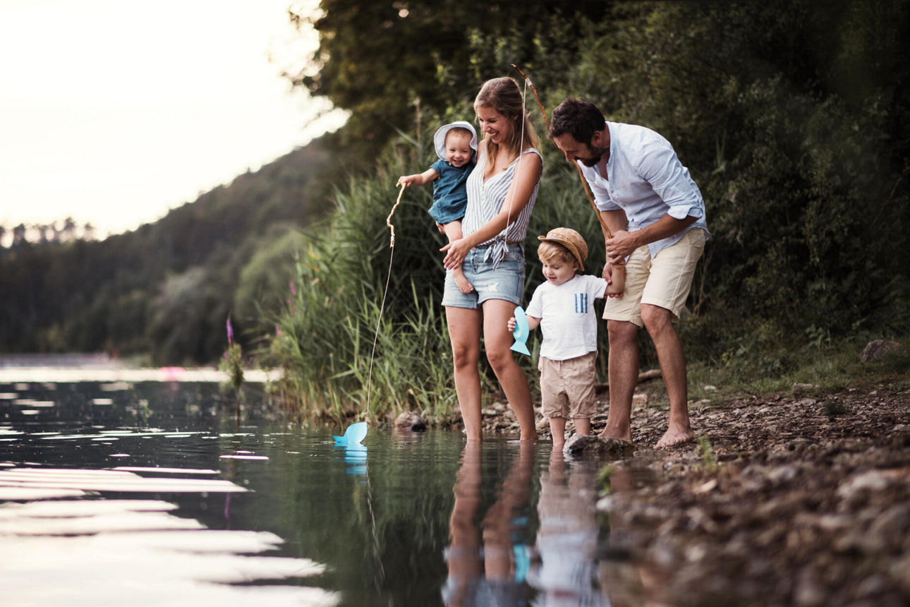 Eine junge Familie steht barfuß an einem See, manche haben die Füße im Wasser