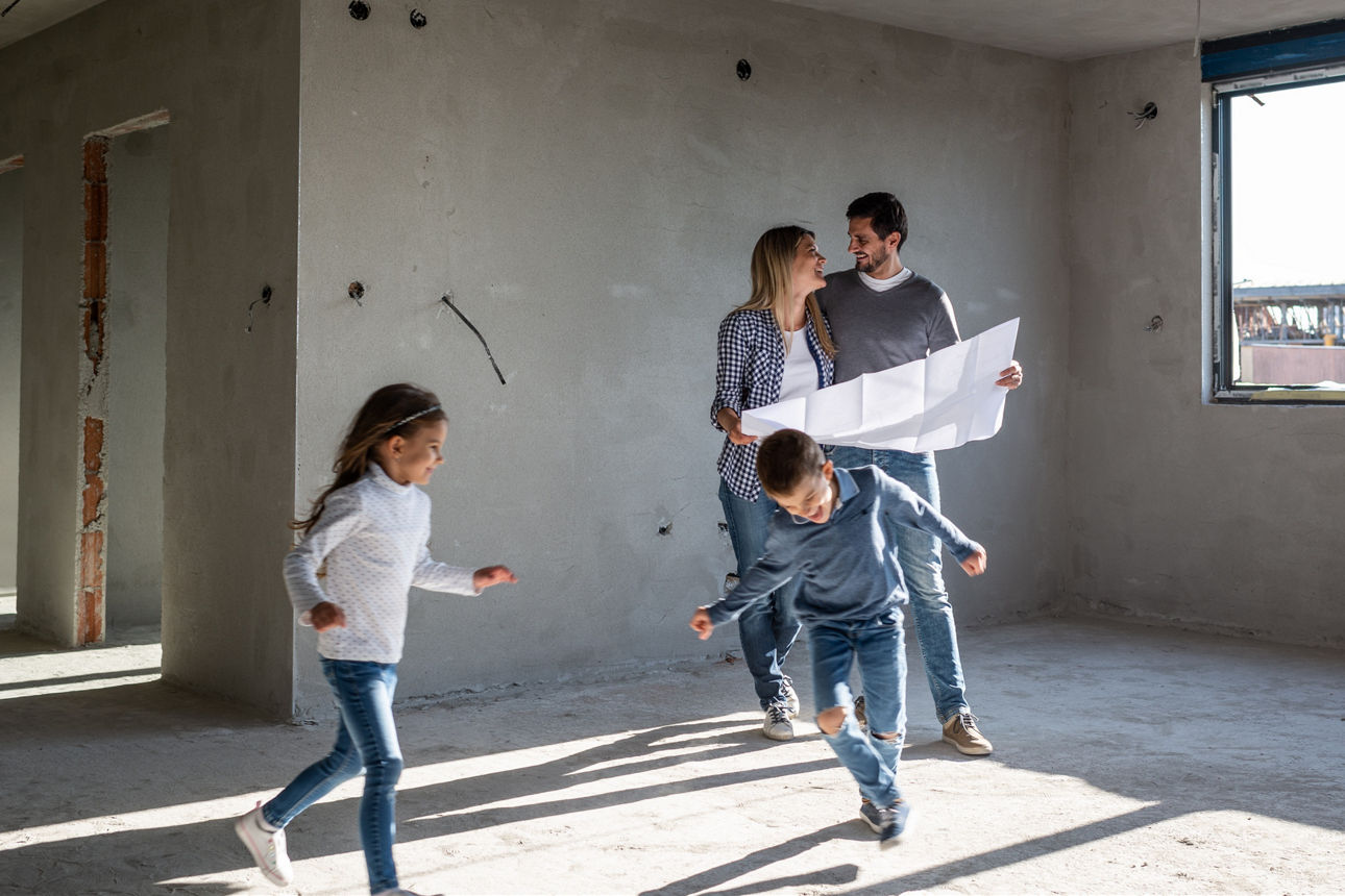 In der Mitte steht ein Pärchen in einem Rohbau. Es fehlen noch Fenster und Türen. Um sie herum rennen zwei Kinder, ein Junge und ein Mädchen. Die Eltern halten einen Plan in der Hand und lächeln sich an.