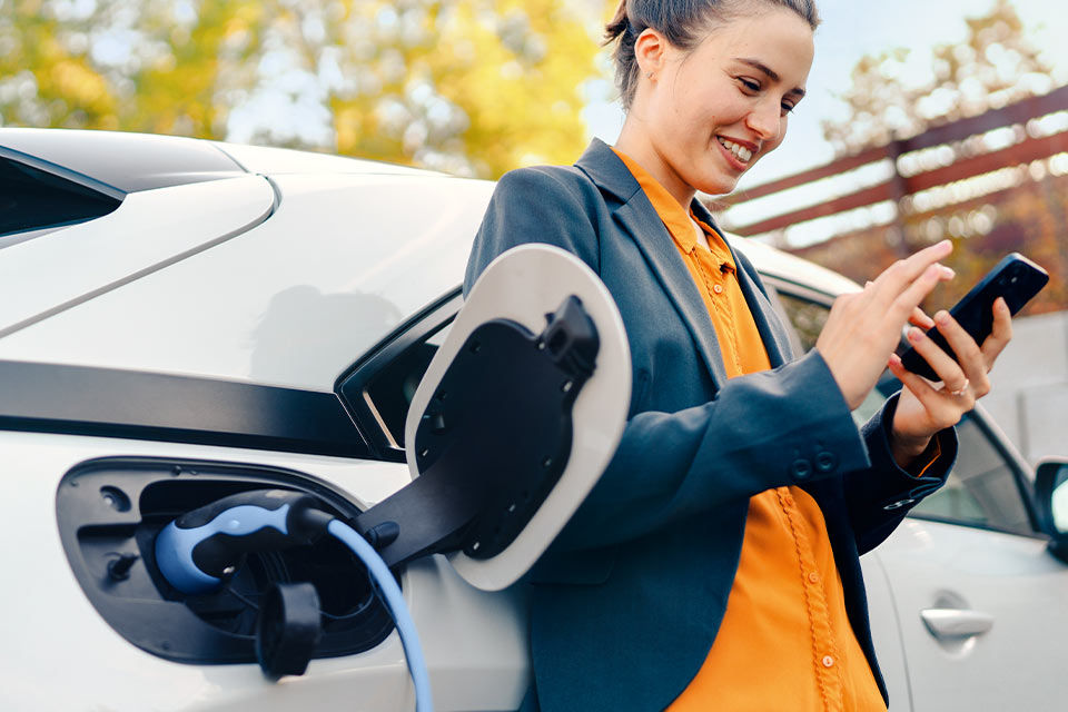 Eine Frau lädt ihr weißes E-Auto draußen auf und hat ein Smartphone in der Hand.
