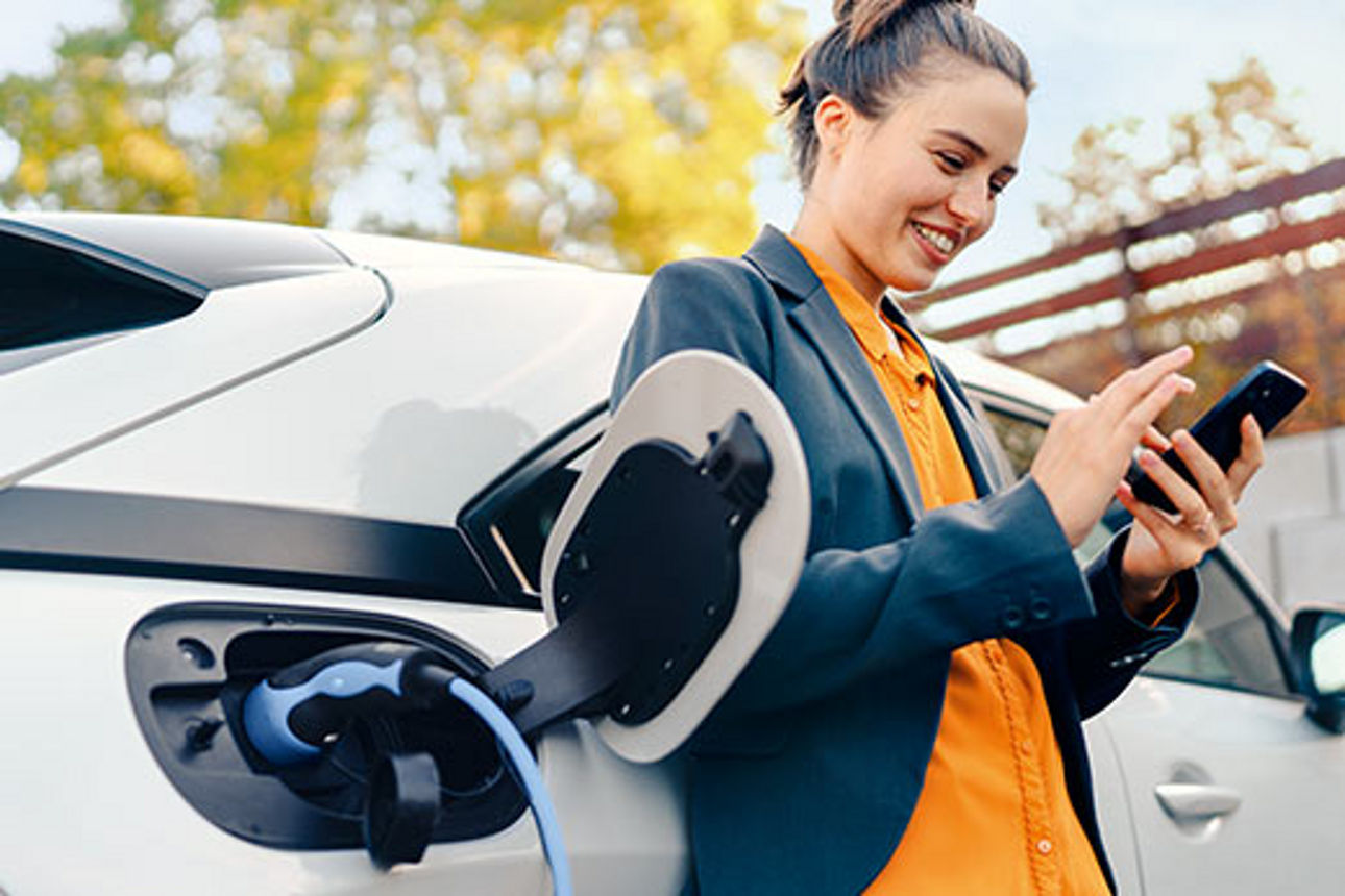 Eine junge Frau lehnt sich beim Tanken an ihr Auto und schaut auf ihr Handy