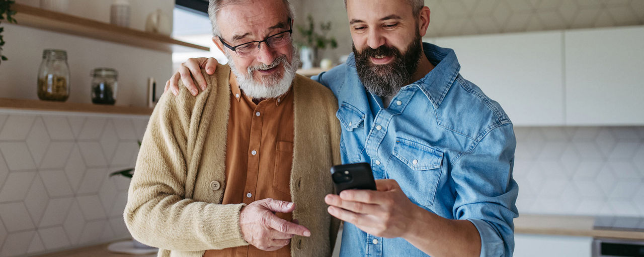 Ein älterer und ein jüngerer Mann schauen in ihrer Küche gemeinsam auf ein Handy
