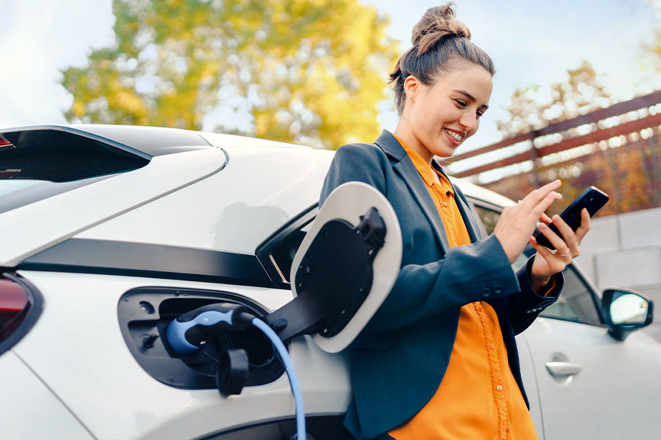 Frau steht angeleht am ladenen E-Auto mit einem Handy in der Hand