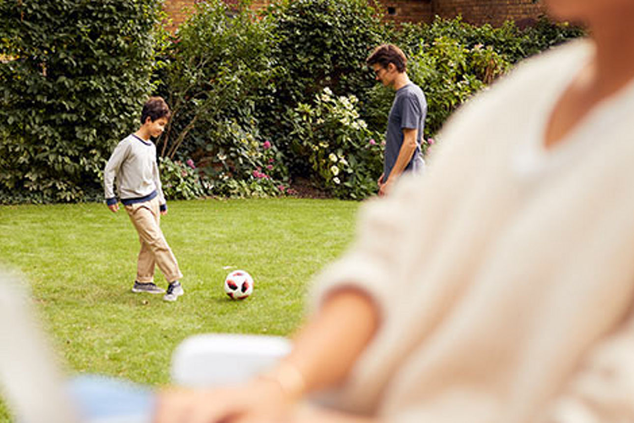 Eine Frau sitzt mit Laptop im Garten, während ihr Sohn und ihr Mann Fußball spielen