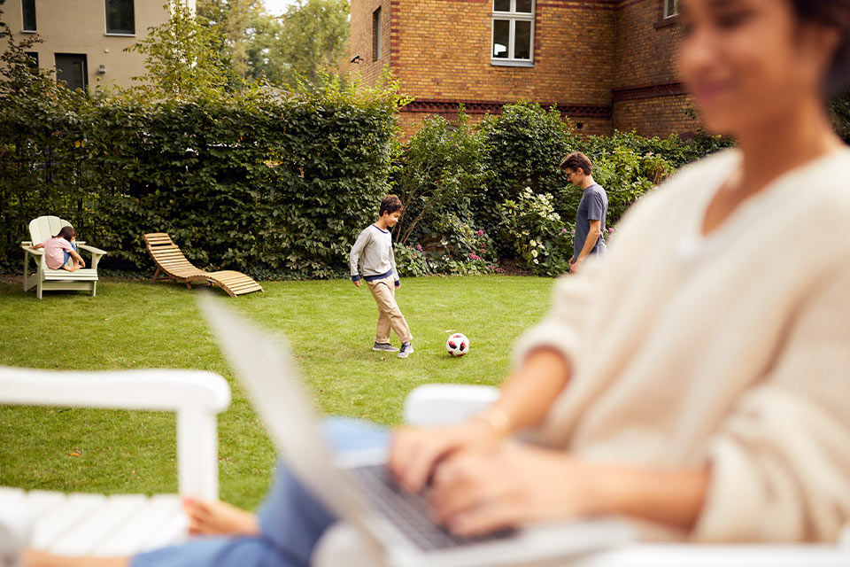 Eine Frau sitzt mit Laptop im Garten, während ihr Sohn und ihr Mann Fußball spielen