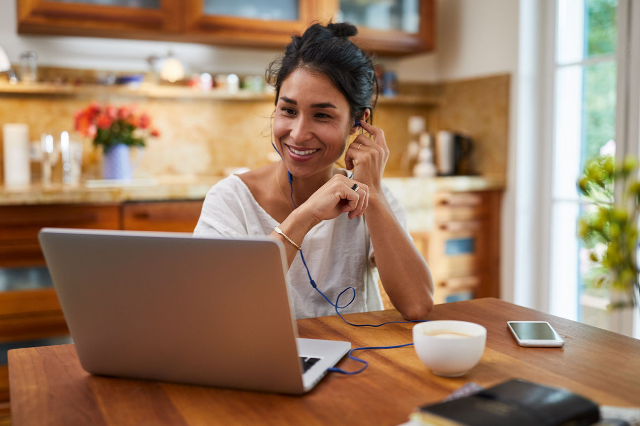 Junge Frau mit dunklen Haaren sitzt mit Kopfhörern vor einem Laptop an einem Holztisch