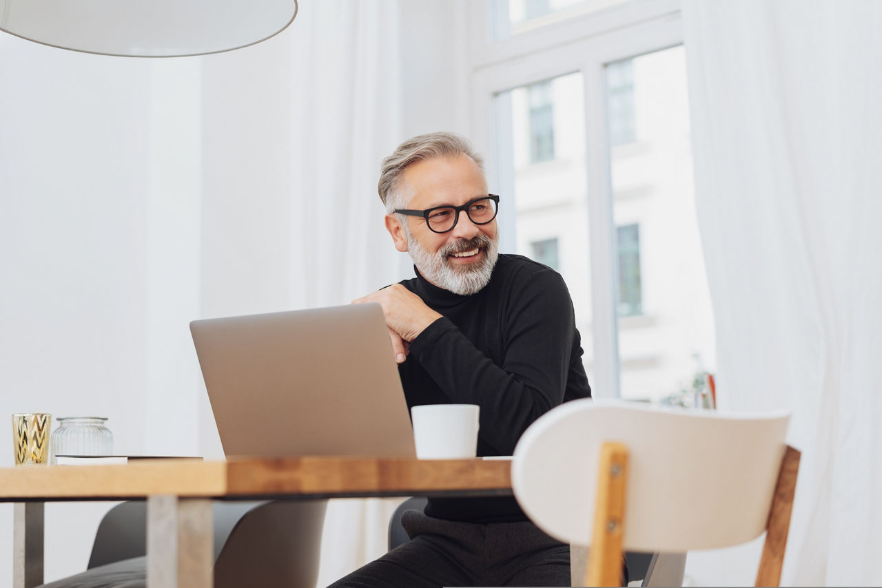 Business Mann sitzt zu Hause am Schreibtisch vor seinem Laptop, schaut lächelnd zur Seite und überlegt wie er sein Geld anlegen kann