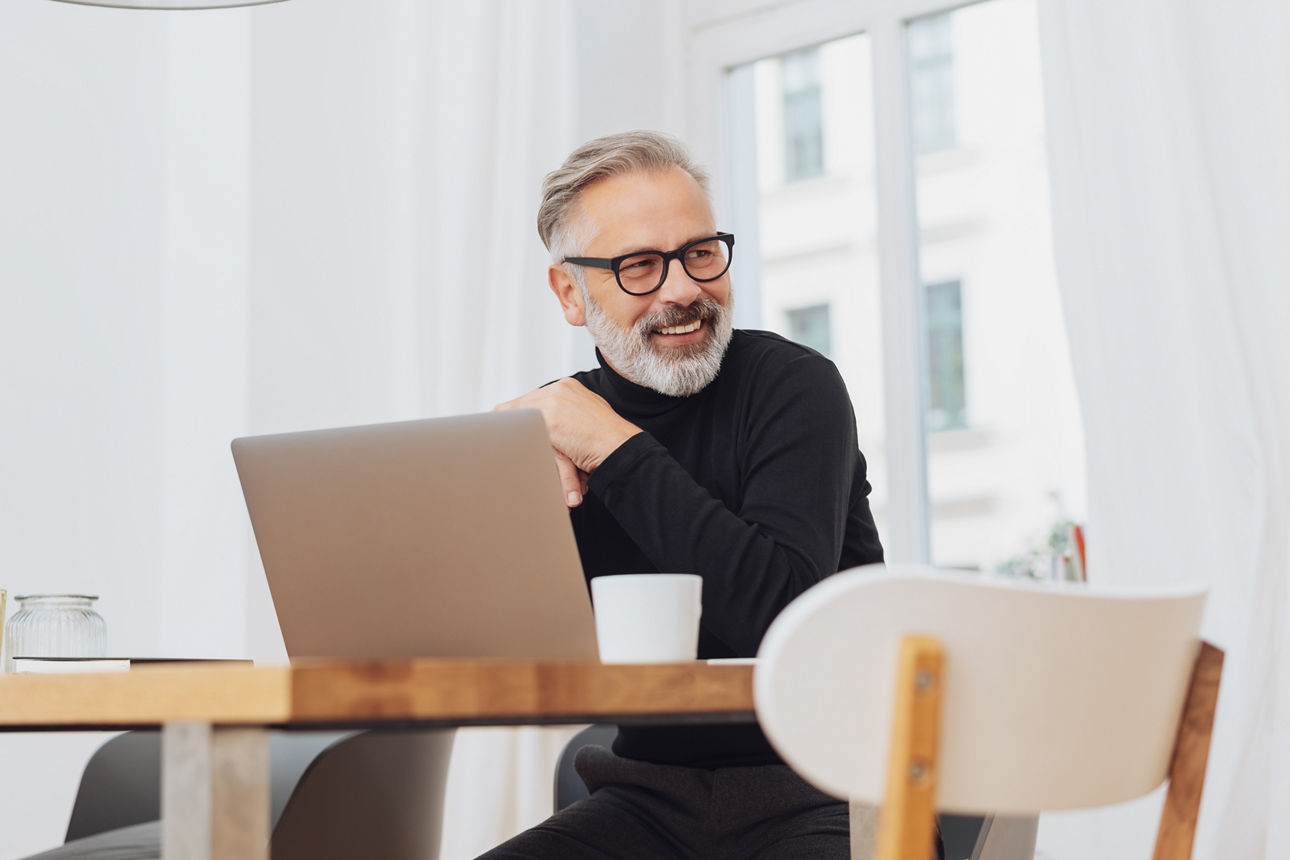 Business Mann sitzt zu Hause am Schreibtisch vor seinem Laptop, schaut lächelnd zur Seite und überlegt wie er sein Geld anlegen kann