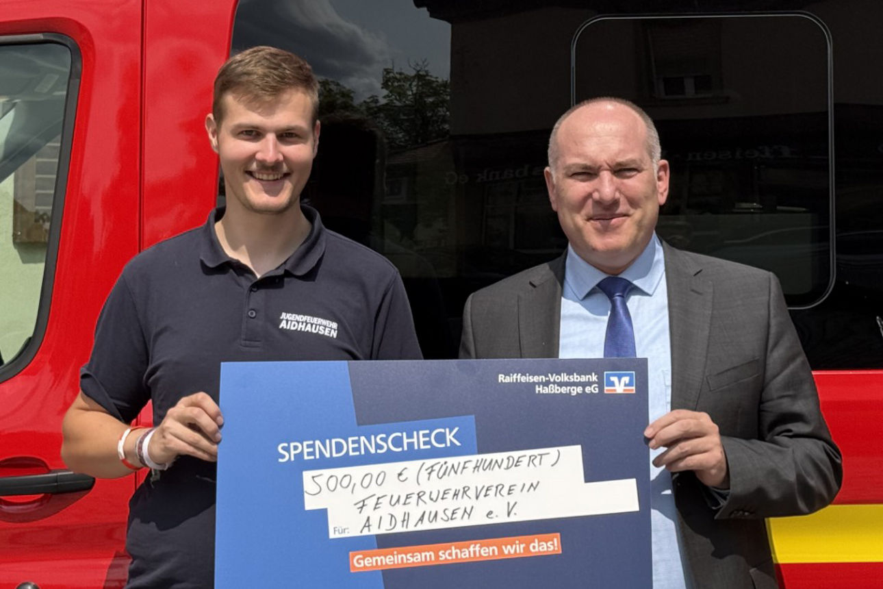 Zwei Männer stehen lächelnd vor einem Feuerwehrauto und halten einen Spendenscheck in ihren Händen