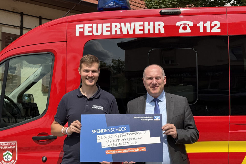Zwei Männer stehen lächelnd vor einem Feuerwehrauto und halten einen Spendenscheck in ihren Händen