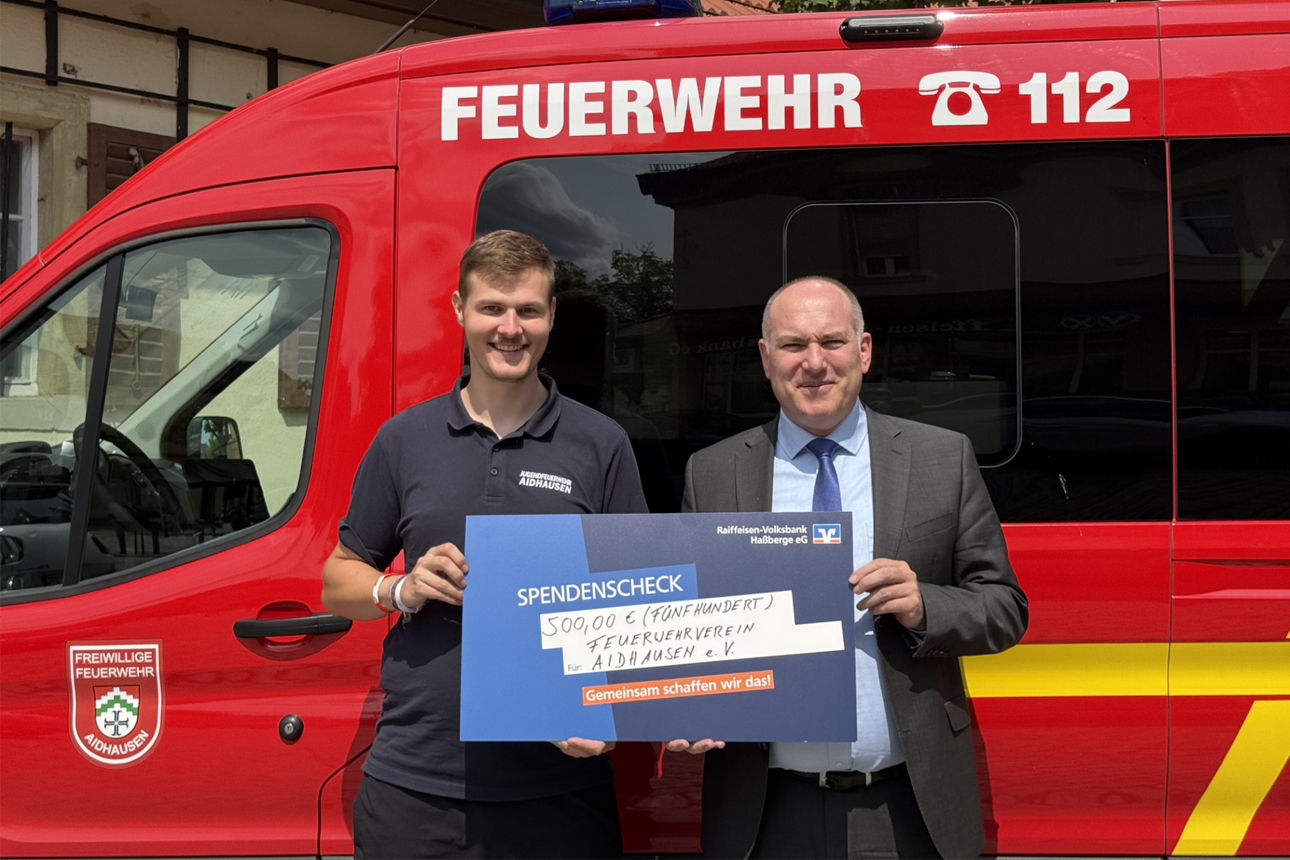 Zwei Männer stehen lächelnd vor einem Feuerwehrauto und halten einen Spendenscheck in ihren Händen