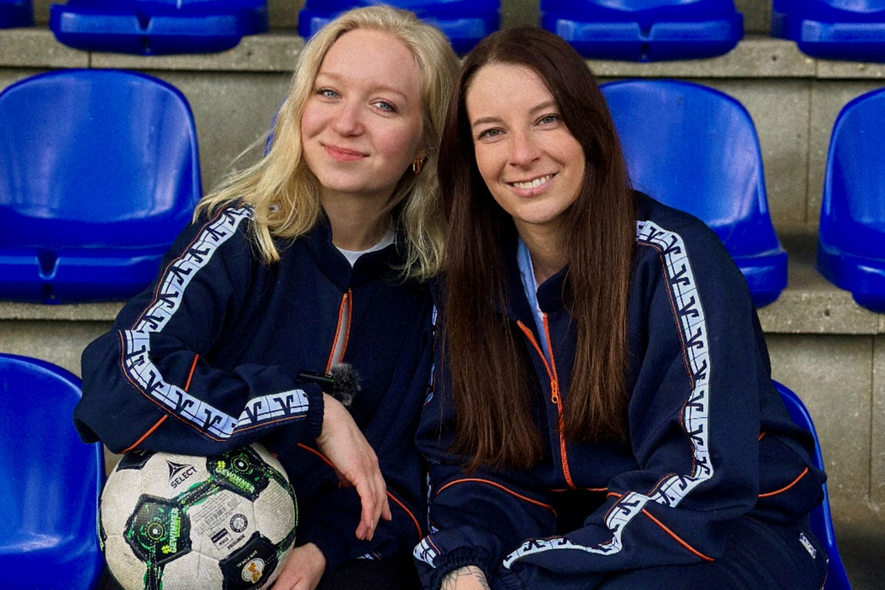 Zwei Frauen sitzen lässig im Zuschauerbereich eines Fußballstadions.