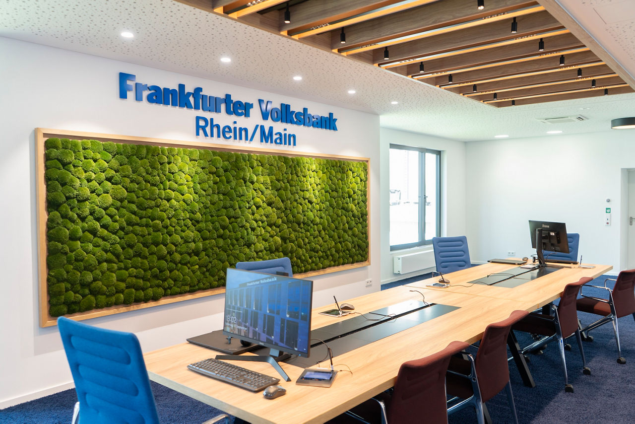 Langer Besprechungstisch mit PCs, im Hintergrund eine Mooswand mit Frankfurter Volksbank Logo darüber