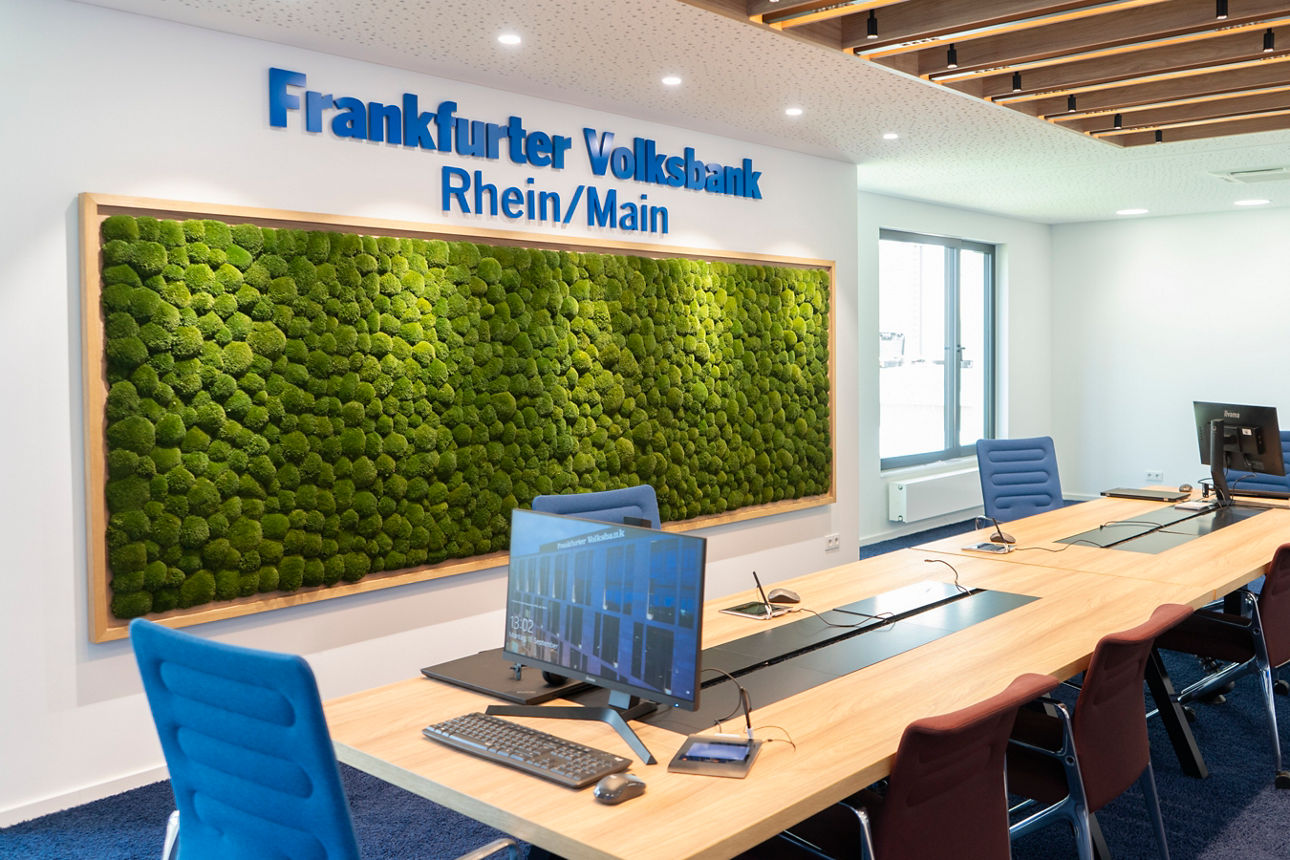 Langer Besprechungstisch mit PCs, im Hintergrund eine Mooswand mit Frankfurter Volksbank Logo darüber