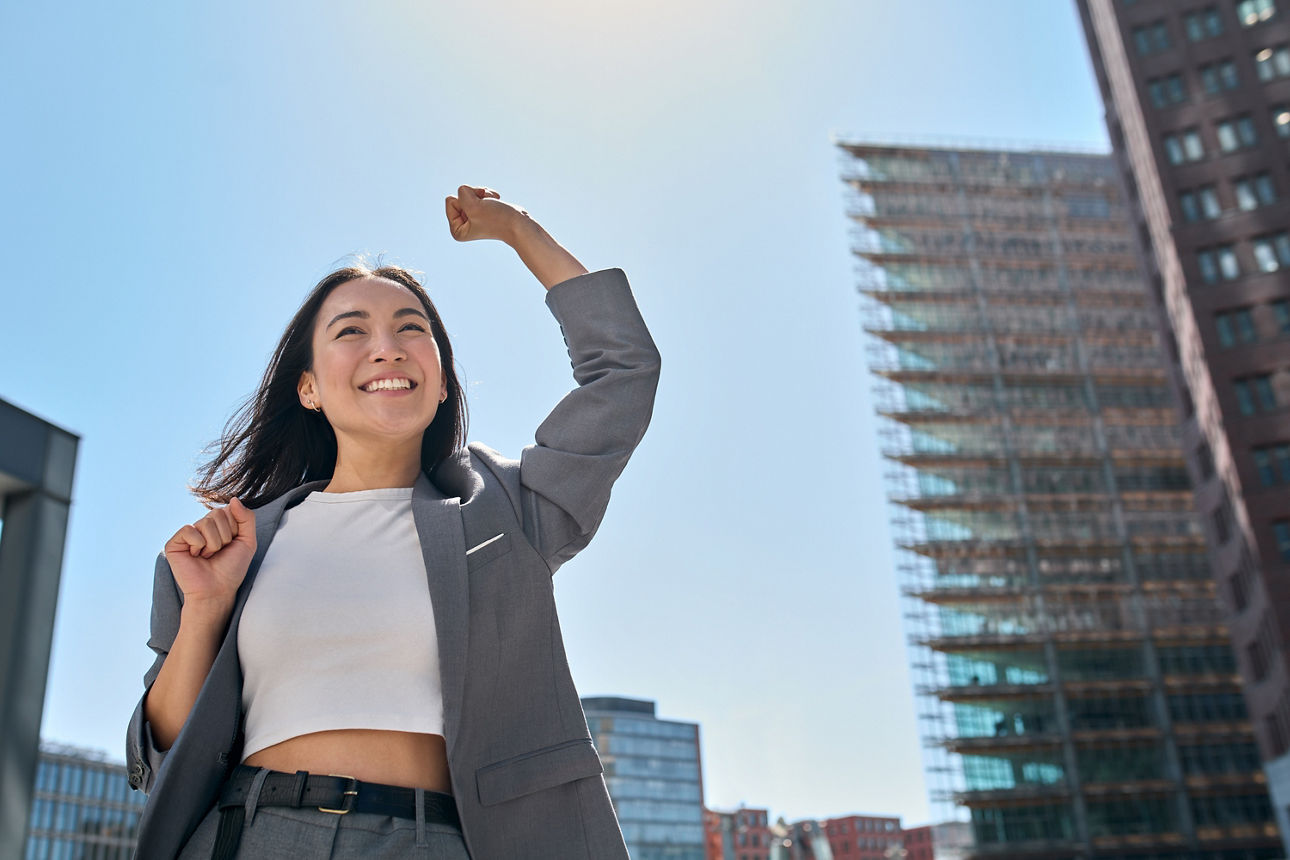 Frau mit erhobenem Arm, freut sich, steht vor einer Skyline unter blauem Himmel