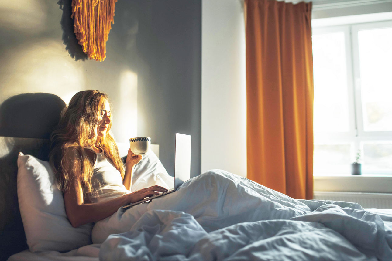 Eine Frau liegt mit einer Tasse Kaffee im Bett und arbeitet am Laptop