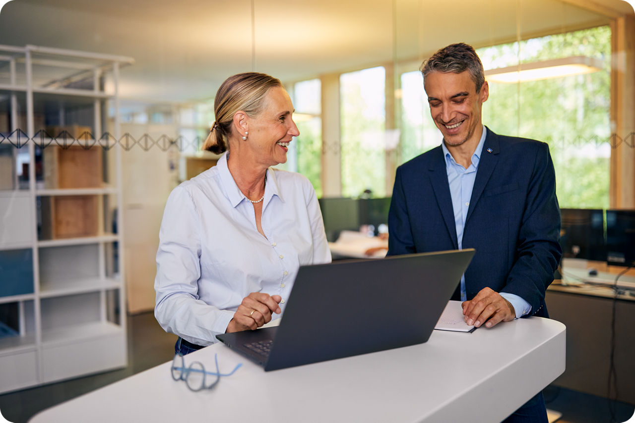 Eine Frau und ein Mann stehen am Tisch und schauen gemeinsam auf ein Notebook