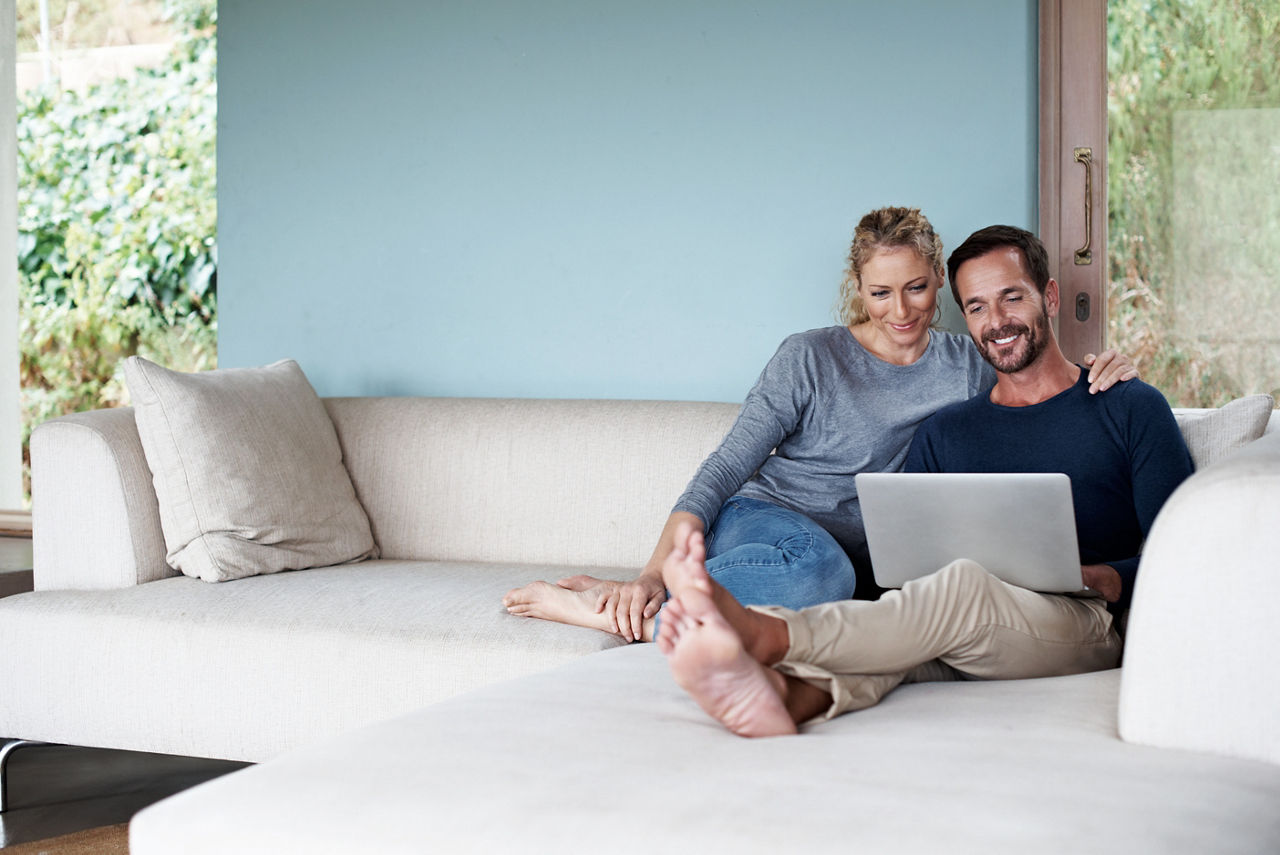 Pärchen liegt gemeinsam mit Laptop auf dem Schoß auf der Couch und unterhält sich über ihre Finanzen