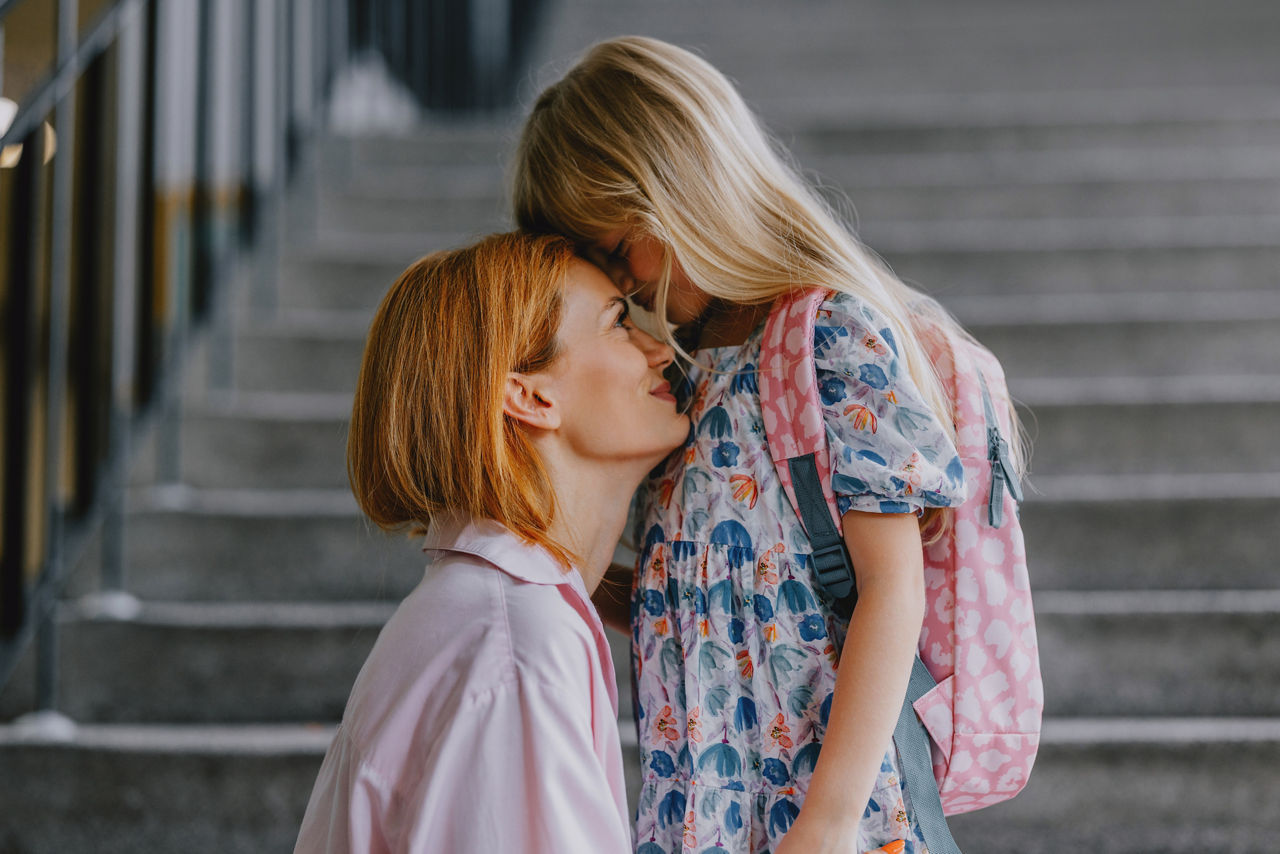 A loving mother kneels to comfort her daughter on the first day of school. The child wears a pink backpack.