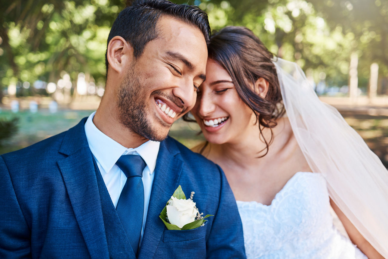 Joyful bride and groom leaning against each other while enjoying romantic moments on wedding day in park on a sunny day