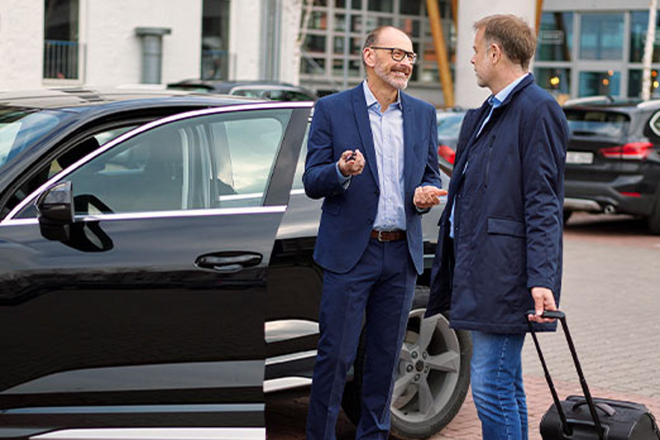 Zwei Männer im Anzug unterhalten sich auf einem Parkplatz vor einer geöffneten Autotür