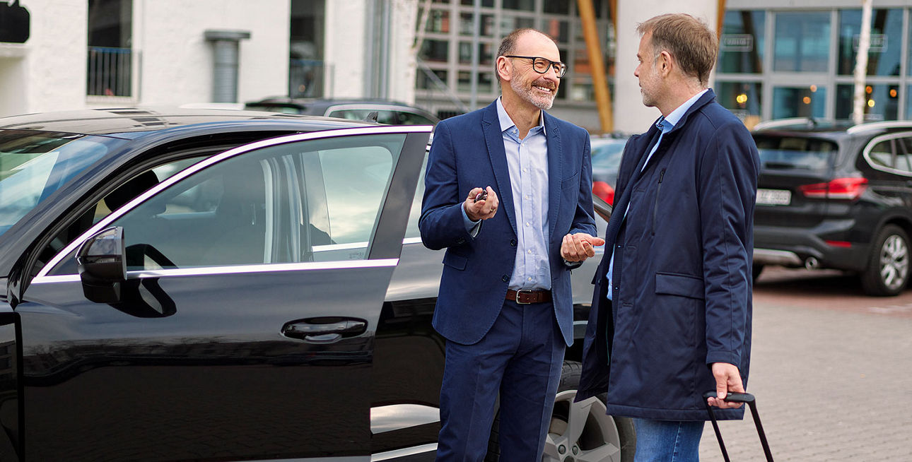 Zwei Männer im Anzug unterhalten sich auf einem Parkplatz vor einer geöffneten Autotür