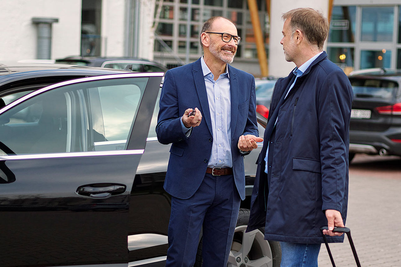 Zwei Männer im Anzug unterhalten sich auf einem Parkplatz vor einer geöffneten Autotür