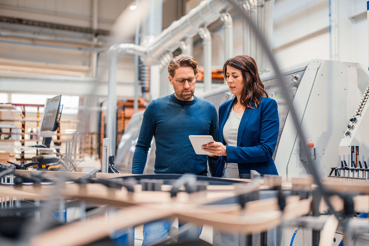Mann und Frau stehen in einer Produktionshalle und schauen auf ein Tablet.