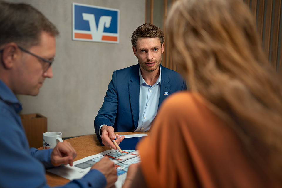 Drei Menschen schauen gemeinsam auf eine Skizze, dahinter das Logo der Volksbanken Raiffeisenbanken