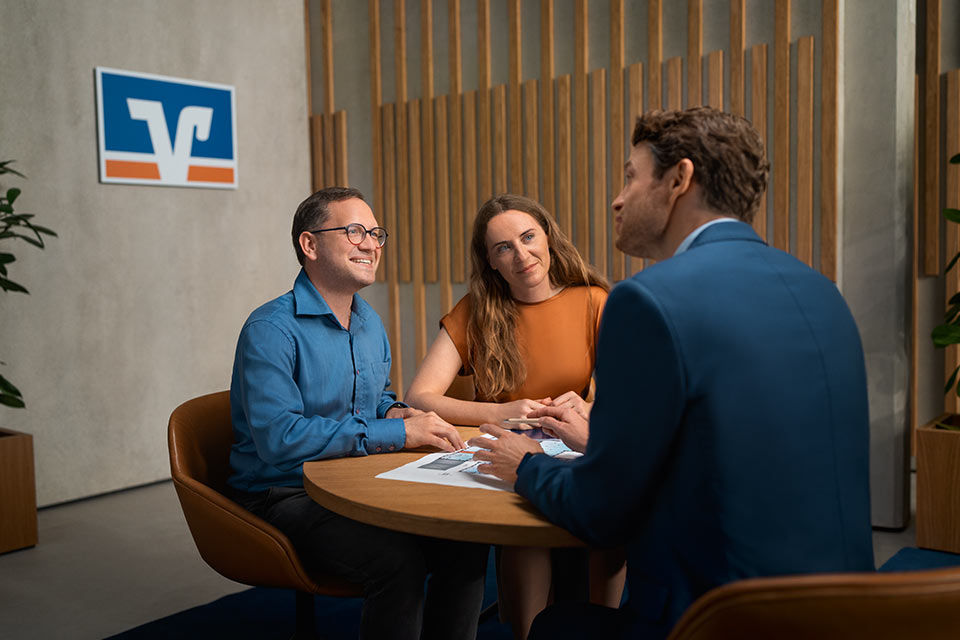 Drei Menschen im Beratungsgespräch, dahinter das Logo der Volksbanken Raiffeisenbanken