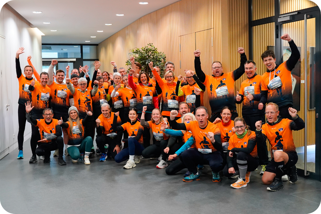 Große Gruppe Teilnehmender in orangefarbenen Laufshirts steht gemeinsam in einem Gebäude und hebt die Arme.
