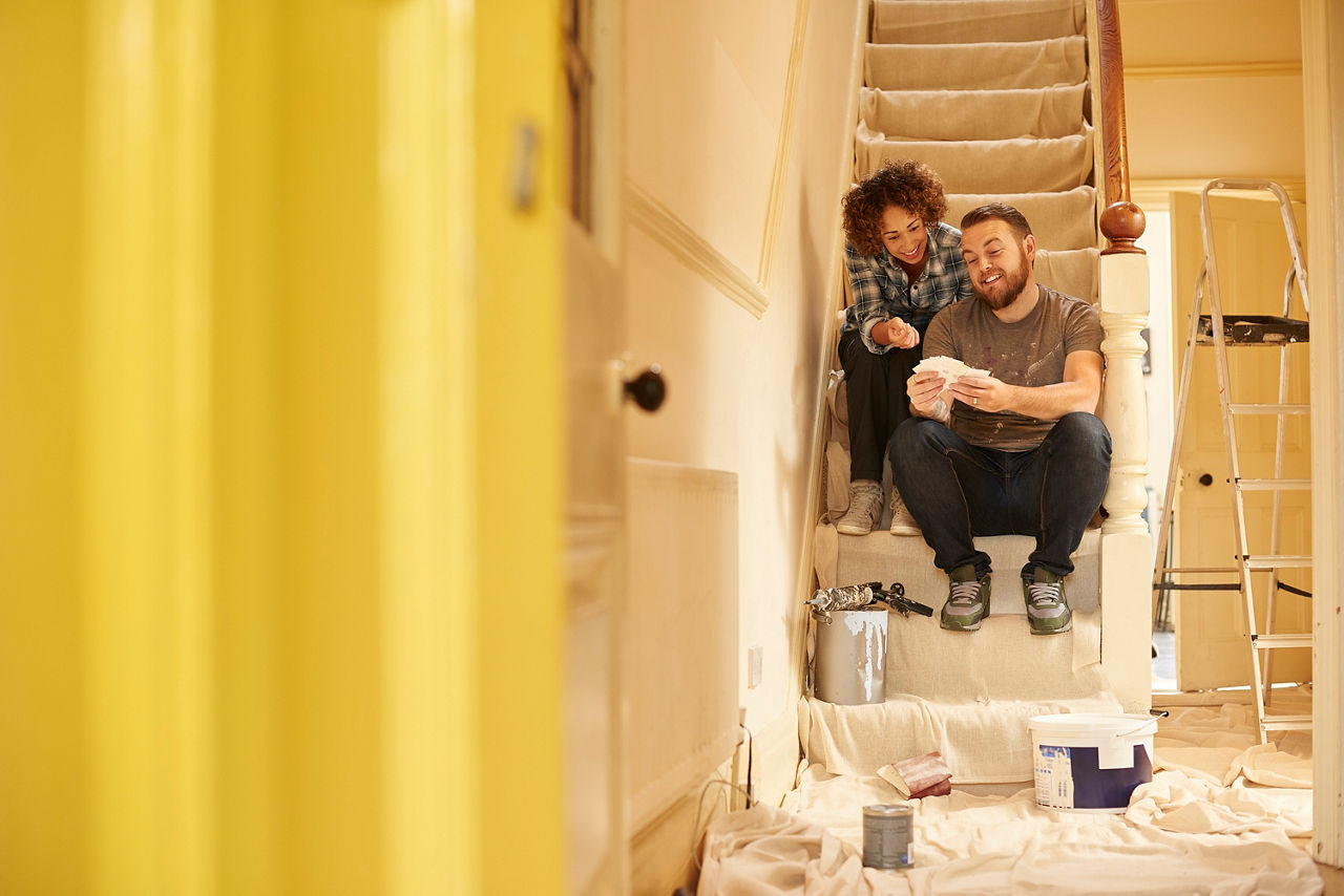 Ein Paar sitzt in einem Haus, welches eine Baustelle ist, im Eingangsbereich auf der Treppe nach oben. Sie sitzt hinter ihm. Er zeigt Ihr eine Auswahl an möglichen Farben für den Gang. 