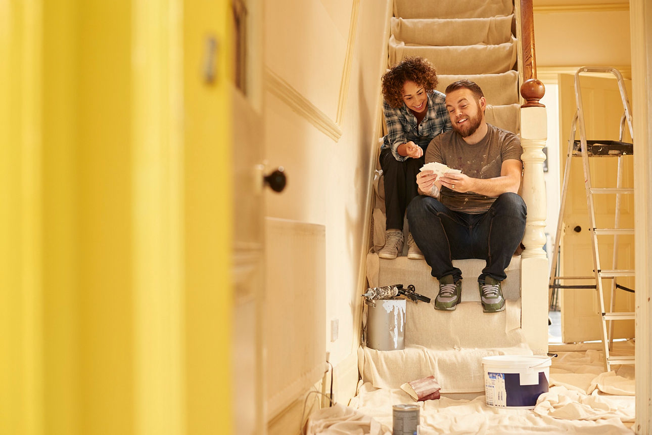 Ein Paar sitzt in einem Haus, welches eine Baustelle ist, im Eingangsbereich auf der Treppe nach oben. Sie sitzt hinter ihm. Er zeigt Ihr eine Auswahl an möglichen Farben für den Gang. 