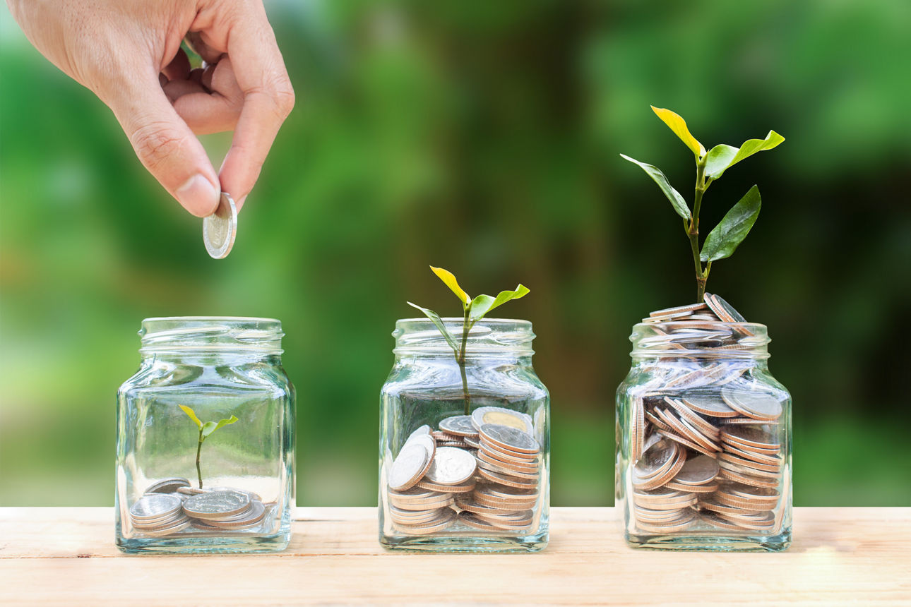 Geld sparen, investieren, Geld für die Zukunft verdienen, Konzept zur finanziellen Vermögensverwaltung. Eine Männerhand, die eine Münze über gestapelte Münzen in einem Glasgefäß und eine wachsende Baumpflanze hält, stellt Fondswachstum und Reichtum dar